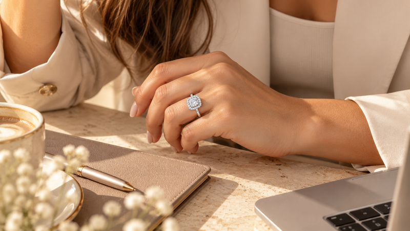 Cushion halo diamond engagement ring worn on a woman's hand in a warm, modern office setting with a cream outfit and natural sunlight.