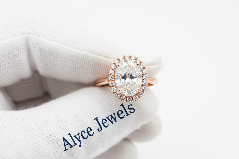 8K close-up of a rose gold oval halo ring held with white cotton gloves featuring a deep blue serif ‘Alyce Jewels’ print. Bright halo details, clean reflections