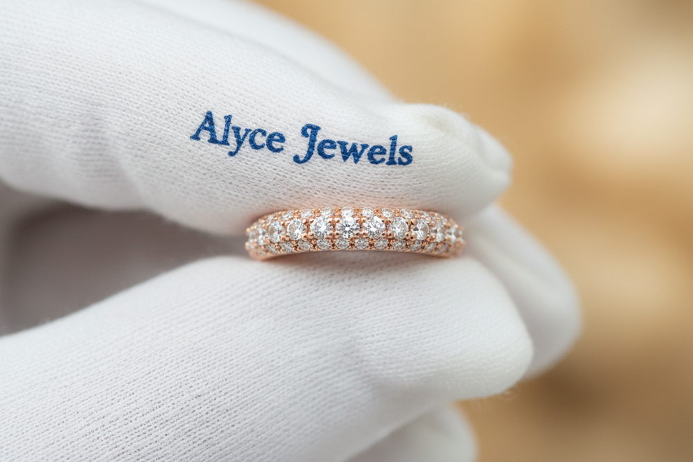 8K close-up of a rose gold pavé wedding band held between white gloves with a deep blue ‘Alyce Jewels’ serif print on it. Sharp stone detail and soft shadows. 
