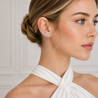 A close-up of a woman wearing a delicate cluster stud earring with sparkling clear stones, styled in a white halter-neck top against a soft, light gray paneled background.