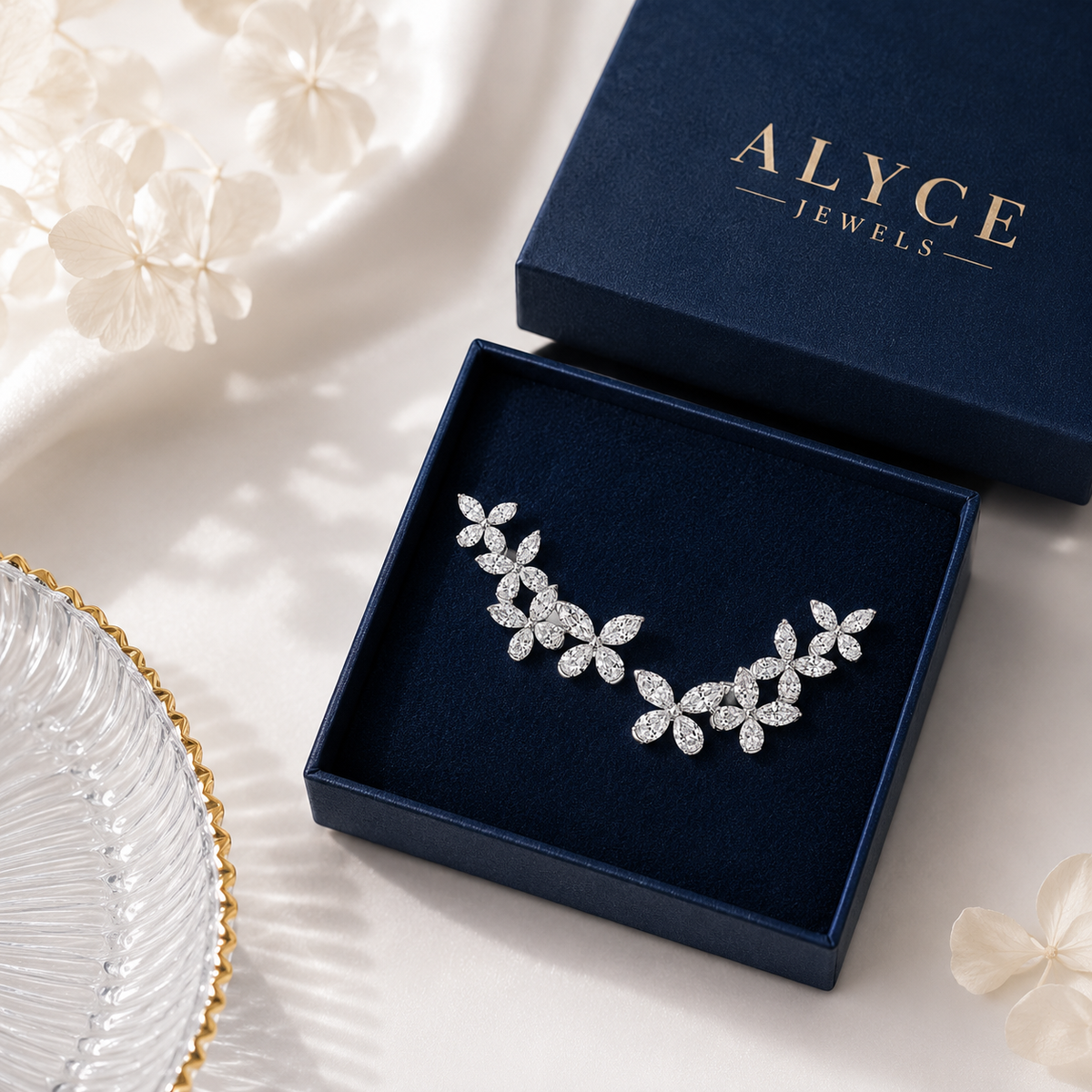 A silver-toned leafy branch hair claw clip set with clear crystals, presented in an open navy blue jewelry box. The navy blue lid of the box is shown behind it with the brand name 'ALYCE JEWELS' in gold lettering.