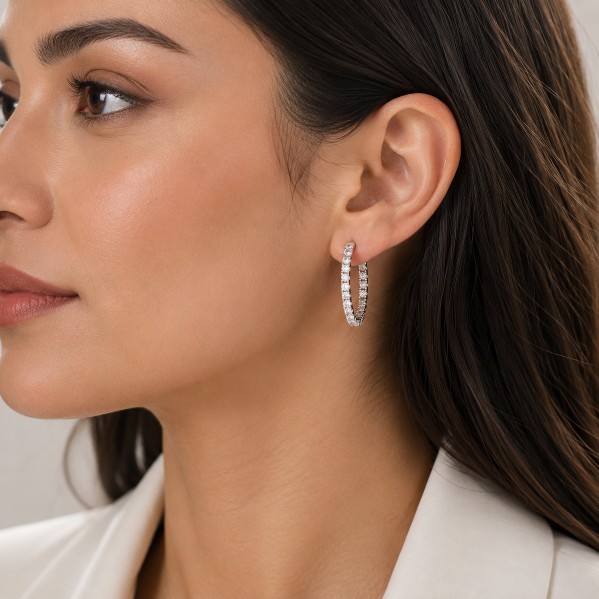 A close-up of a woman wearing a small silver hoop earring set with clear stones. The image focuses on the earring on her ear, with her face and hair blurred out.