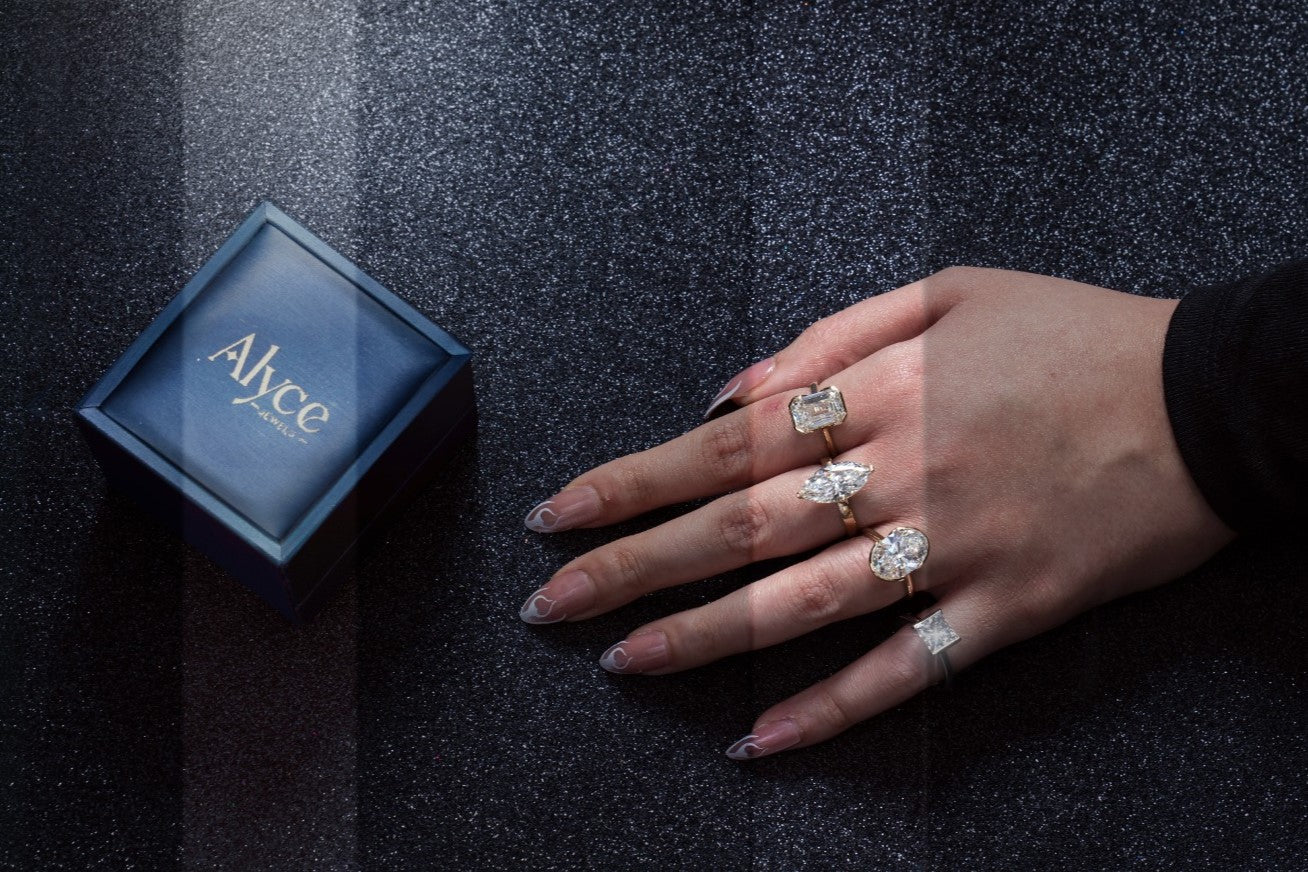 Hand wearing multiple rings with a 'Alyce' jewelry box on a dark textured surface
