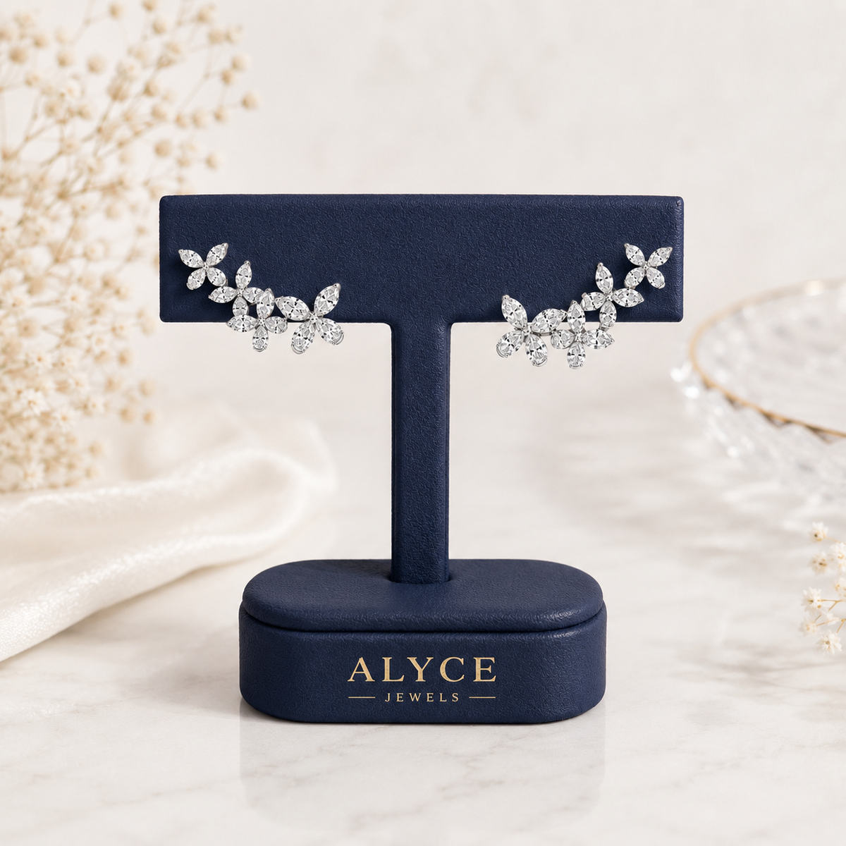 A pair of silver-toned leaf cluster stud earrings set with clear stones, displayed on a navy blue T-stand jewelry holder with the brand name ALYCE JEWELS visible on the base. The setup is styled on a light marble surface with soft decorative elements in the background.