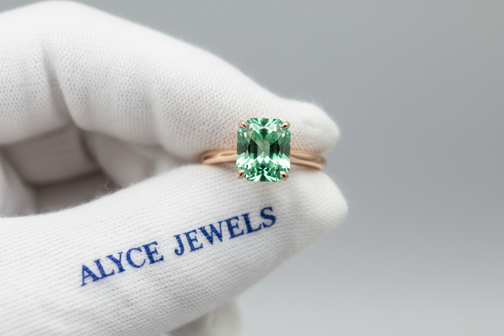 close-up of a rose gold emerald solitaire ring held with white cotton gloves printed with a blue serif ‘Alyce Jewels’ print on it. Strong reflections and crisp facets